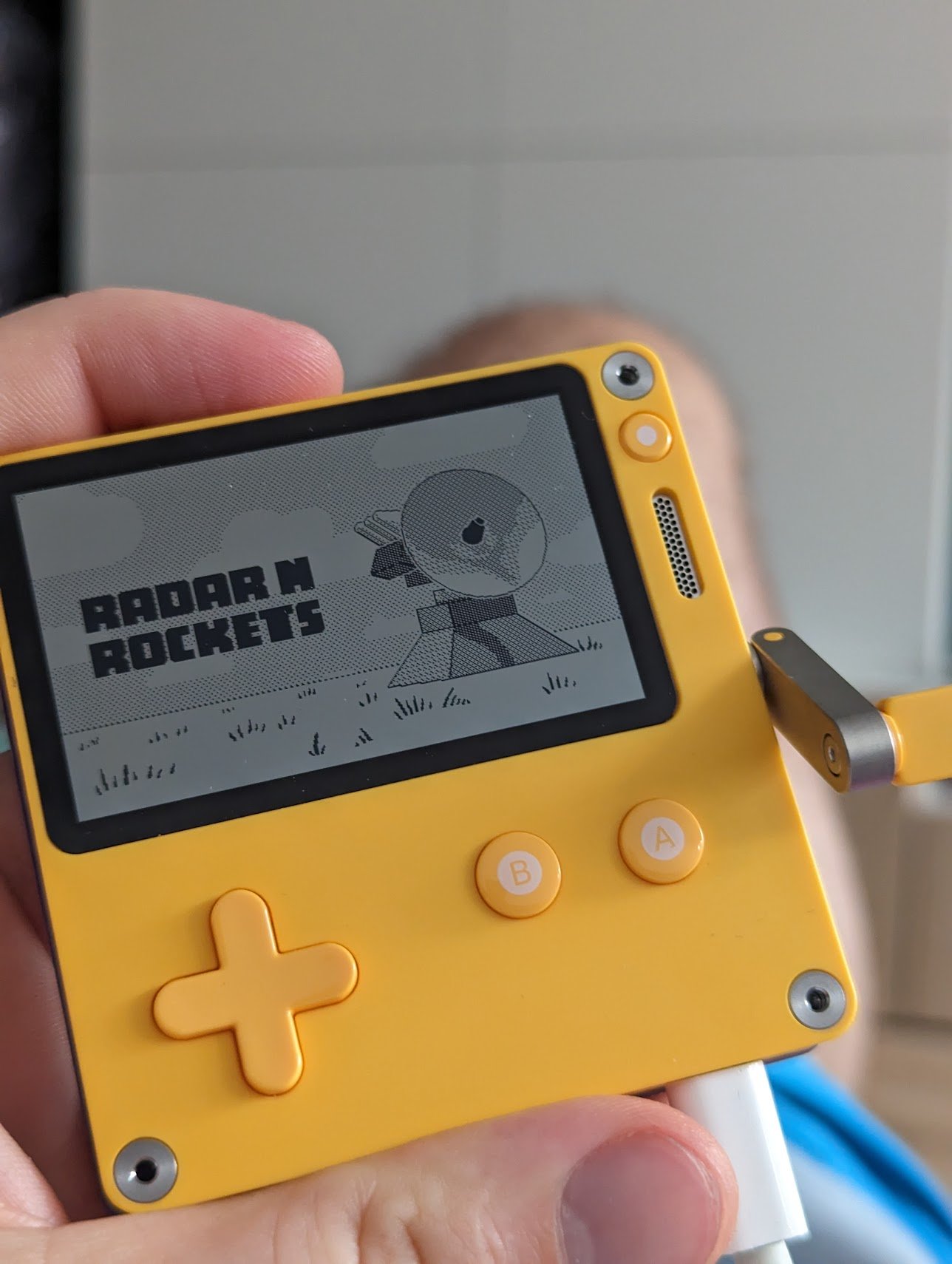 Yellow Playdate in hand showing the Radar N Rockets title screen — a 1-bit radar dish beside the game name.